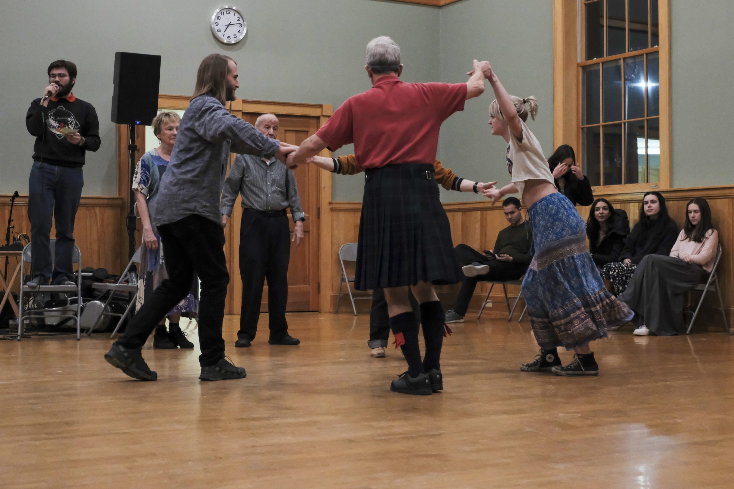 A group of five people of different ages and genders hold hands in a circle and dance. A caller is seen to the left and others are sitting to the right.